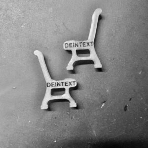 Zwei transparente Miniatur-Parkbänke aus Kunststoff liegen auf einer dunklen Oberfläche. Die Sitzflächen sind mit dem schwarzen Schriftzug „DEINTEXT“ als Platzhalter bedruckt.