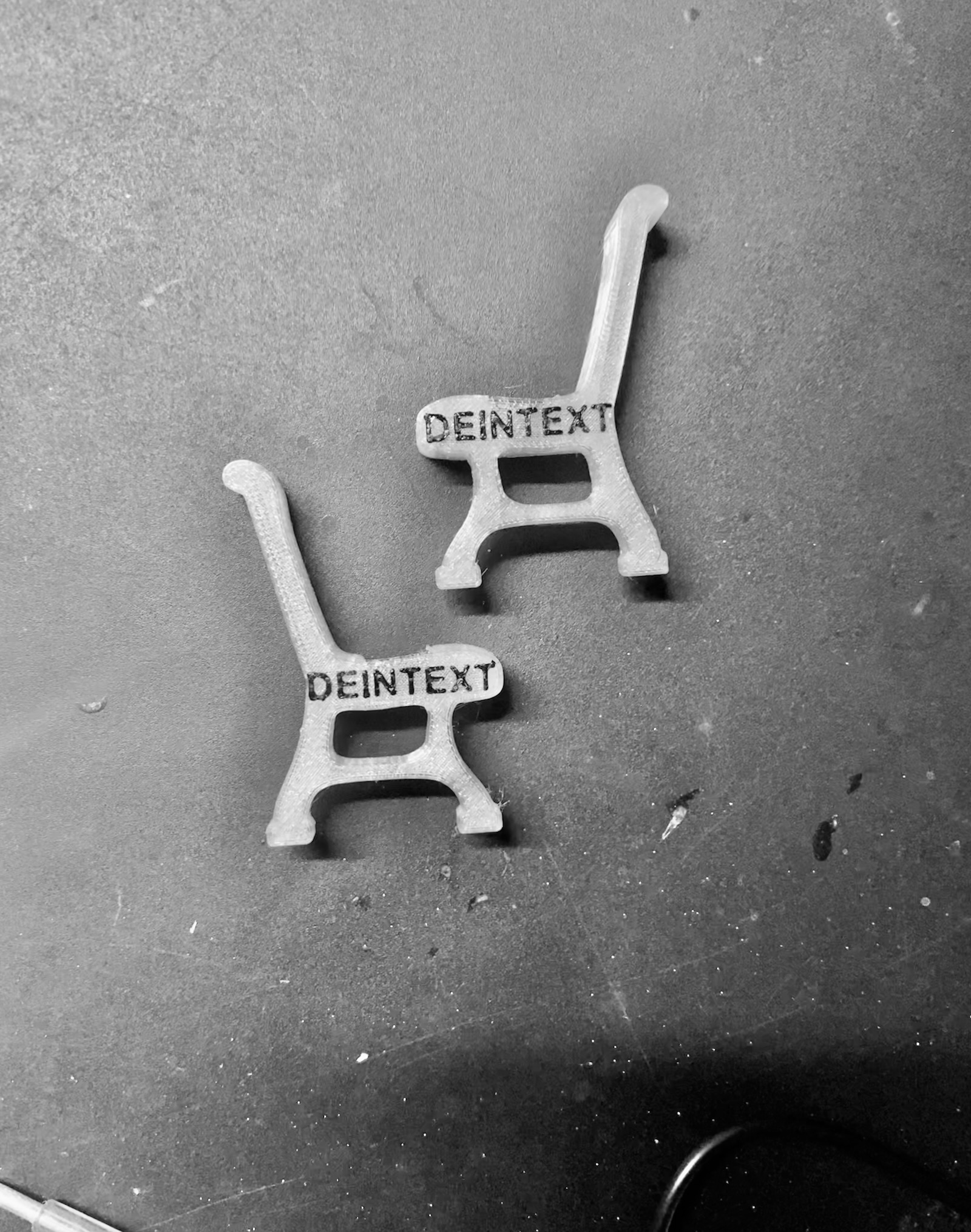 Zwei transparente Miniatur-Parkbänke aus Kunststoff liegen auf einer dunklen Oberfläche. Die Sitzflächen sind mit dem schwarzen Schriftzug „DEINTEXT“ als Platzhalter bedruckt.