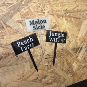Drei rechteckige Pflanzstecker aus Kunststoff auf einer OSB-Holzplatte. Die Schilder sind mit den Begriffen „Melon Sicle“, „Peach Farts“ und „Jungle WIFI“ beschriftet.